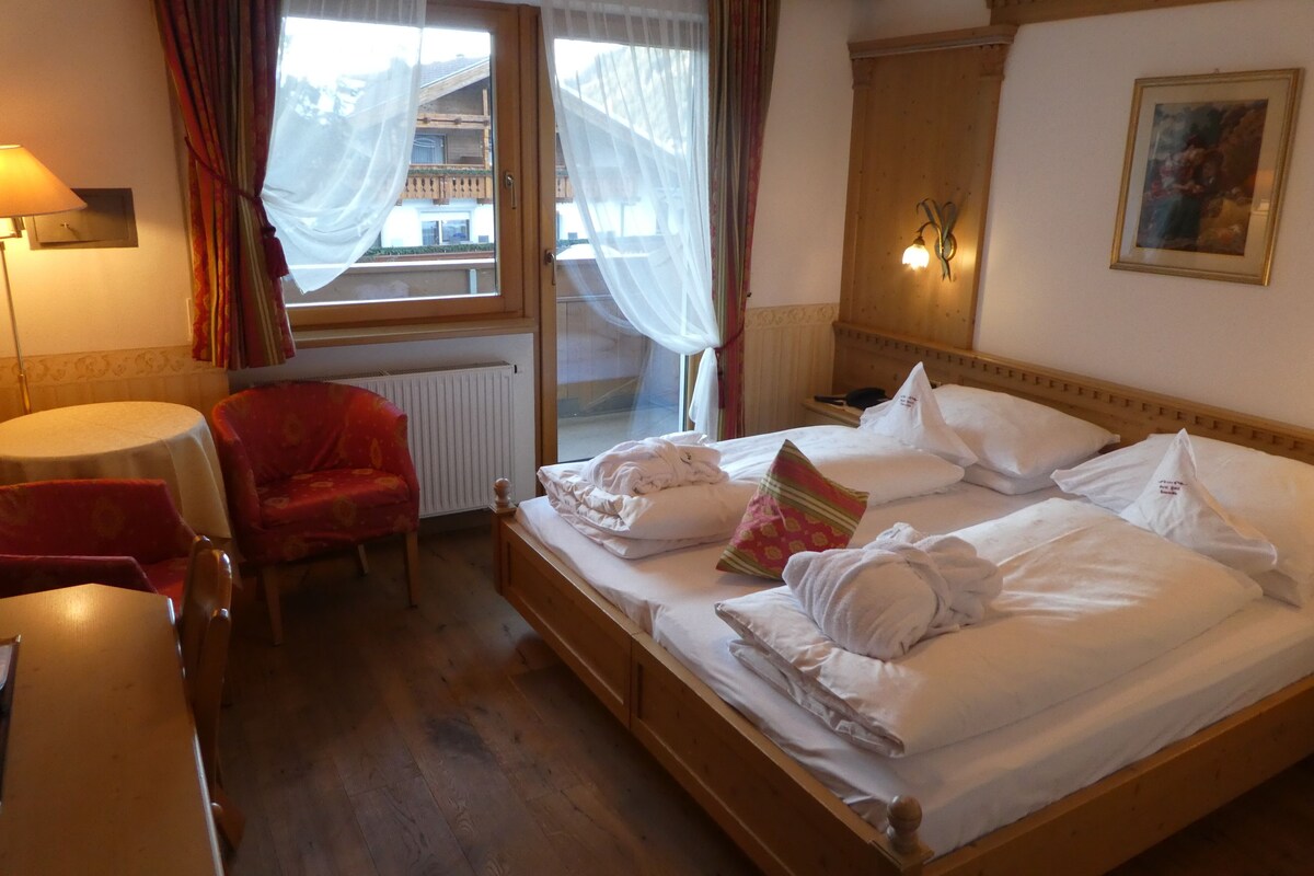 A hotel room with a wooden bed, a red chair, a table, and a window with curtains.