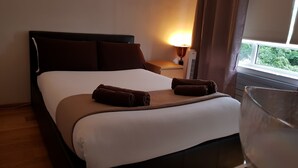 Cama bem arrumada com lençol branco e duas toalhas castanhas, abajur e janela com cortinas.