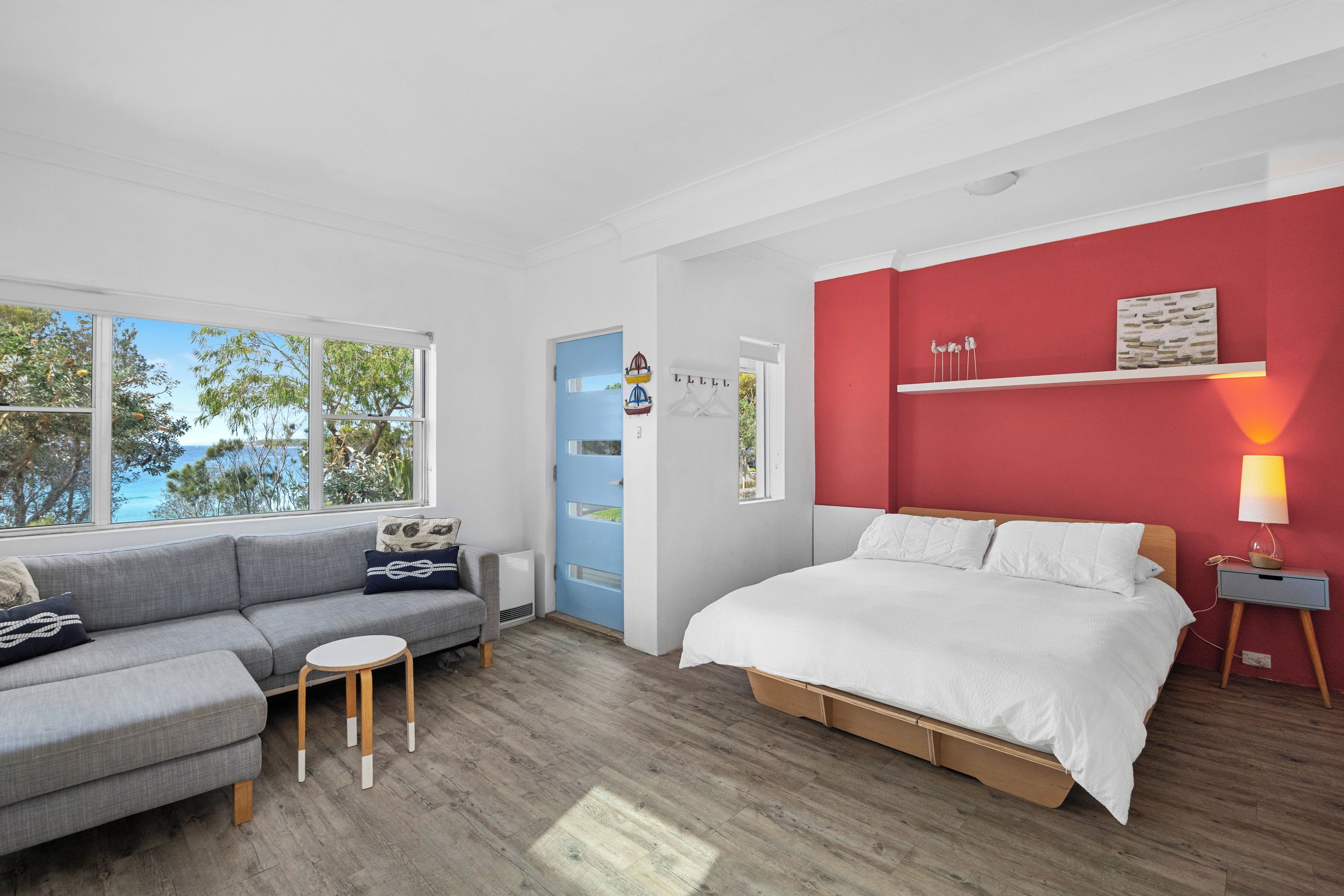 A modern bedroom with a grey sofa, a bed with white bedding, a red accent wall, and a view of trees through the window.