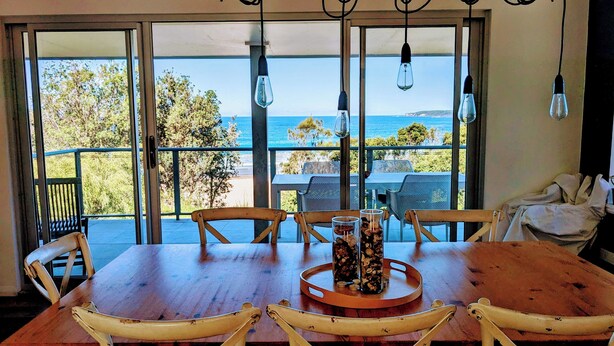 A wooden dining table with four chairs, a glass vase with dried flowers, and a view of the sea through large glass doors. - Holiday house right on Mollymook beach (Mollymook Beach)