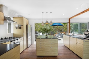 A modern kitchen with wooden cabinets, a central island, and a view of an outdoor patio area. - Beach House - footsteps from the sand (Mollymook Beach)