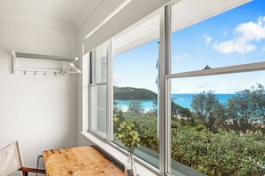 A room with a large window, a wooden table, and a hanging clothes rack. - Beach House - footsteps from the sand (Mollymook Beach)