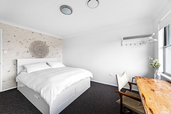 A modern bedroom with a bed, a wooden table, a chair, and a wall-mounted air conditioning unit. - Beach House - footsteps from the sand (Mollymook Beach)