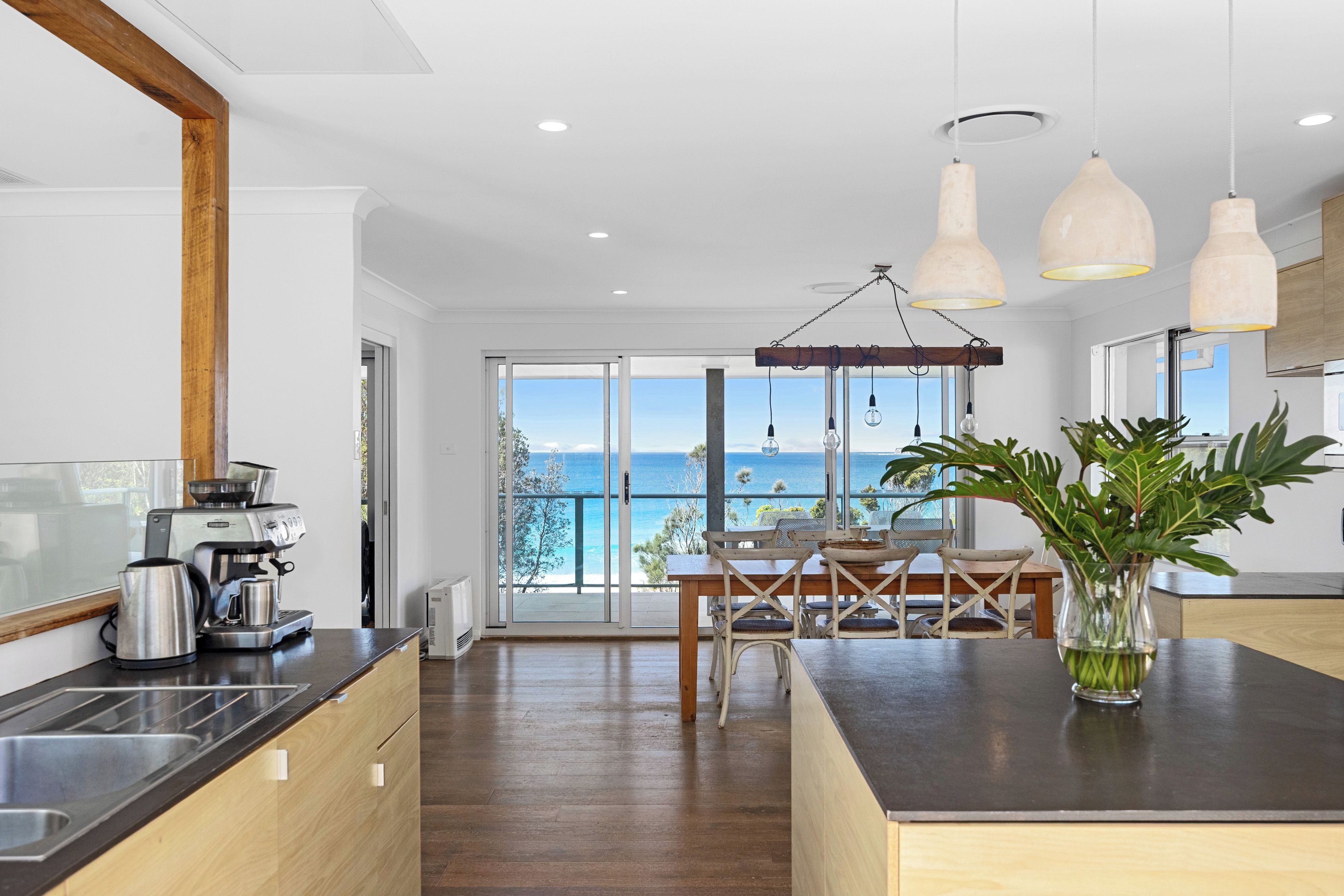 A modern kitchen with a view of the sea, featuring a coffee machine, a sink, and a dining area with a table and chairs.