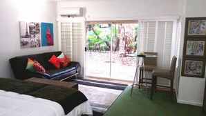 Una habitación con una cama, un sofá de cuero negro con cojines de colores, una mesita con una silla y un ventanal corredero que da a un espacio exterior con plantas.