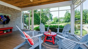 En overdækket udendørs siddeplads med Adirondack-stole og et lille bord, med udsigt over en have og et hus.