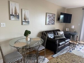 A living room with a glass dining table, black leather sofa, and a flat-screen TV mounted on the wall.