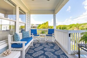 A covered porch with white rocking chairs and blue cushions, a blue patterned rug, and a view of a green landscape.