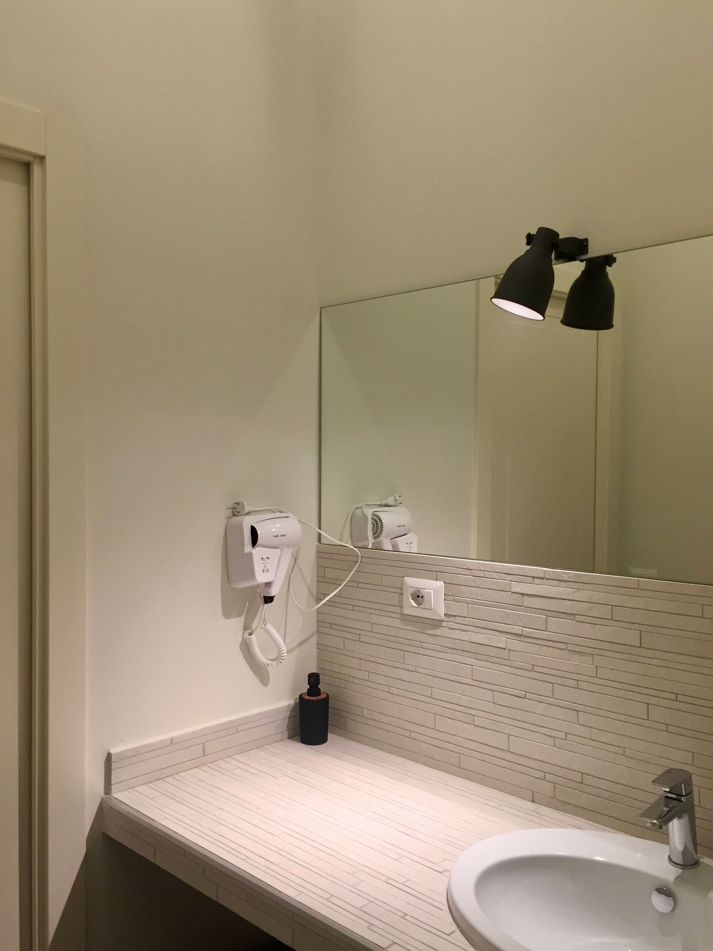 A bathroom with a white sink, a mirror, two hair dryers mounted on the wall, and a black lamp.