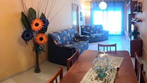 Un salon avec un canapé, une table à manger avec des chaises, un vase de fleurs et un luminaire au plafond.