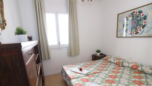 A bedroom with a floral bedspread, a wooden dresser, a small bedside table with a plant, and a window with curtains. - 161 - COSTA AZUL LUXE..Spacious and central apartment with wonderful views (SALOU)