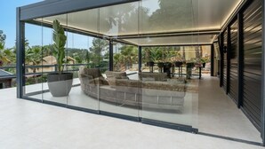 A modern glass-enclosed living area with a sofa and a dining table, surrounded by plants and a view of the outdoors.