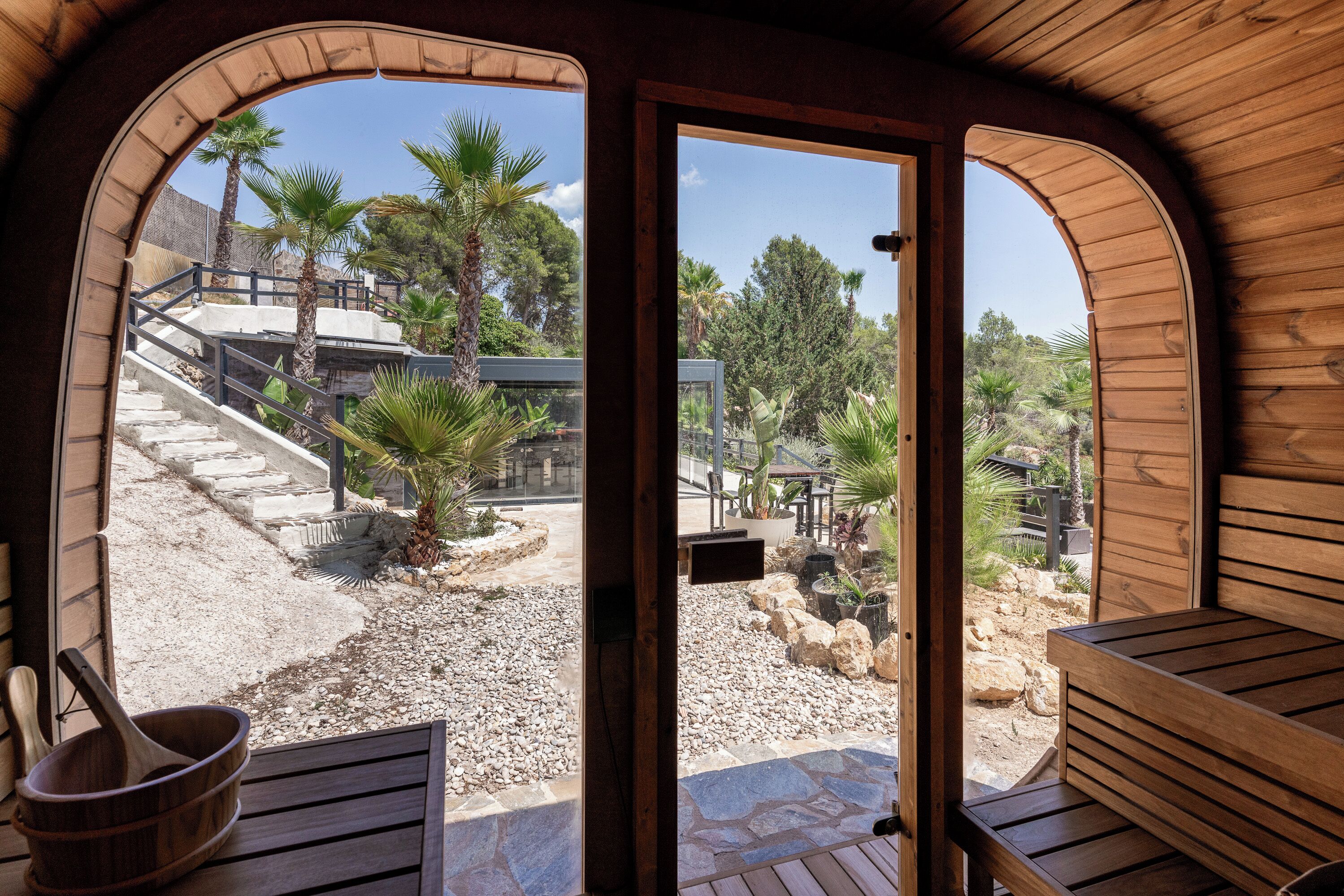 A wooden interior with a view of a landscaped outdoor area including a staircase, palm trees, and a seating area.