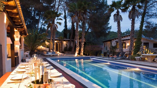 A poolside dining area with tables set for a meal, surrounded by palm trees and a clear pool.