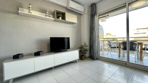 A modern living room with a flat-screen TV on a white cabinet, a potted plant, and a balcony with outdoor furniture.