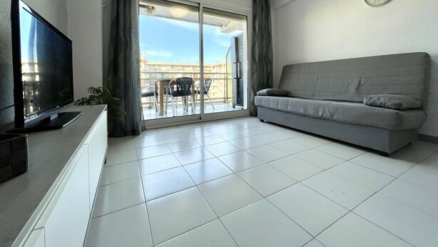 A modern living room with a flat-screen TV, grey sofa, and a sliding glass door leading to an outdoor balcony.