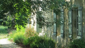 A rustic building with a gravel path leading to a wooden door, surrounded by lush greenery and flowering plants. - Beautiful Stone Farmhouse With Heated Pool And Views Across Rolling Countryside (Gondrin)