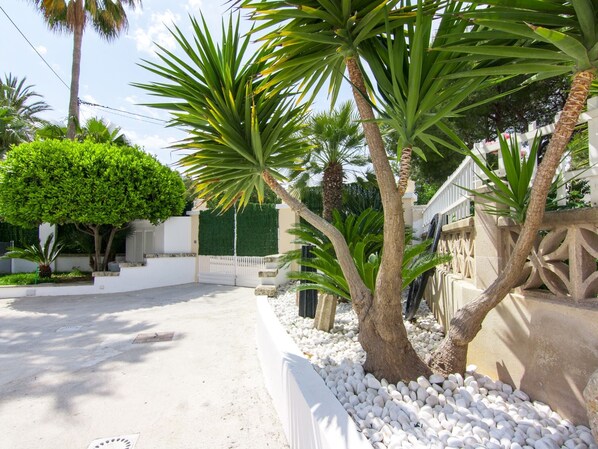 A paved driveway leading to a gated entrance with a white picket fence, surrounded by palm trees and a hedge. - Villa Comes - Beautiful villa with private pool 50 metres from the beach (Port d'Alcúdia)