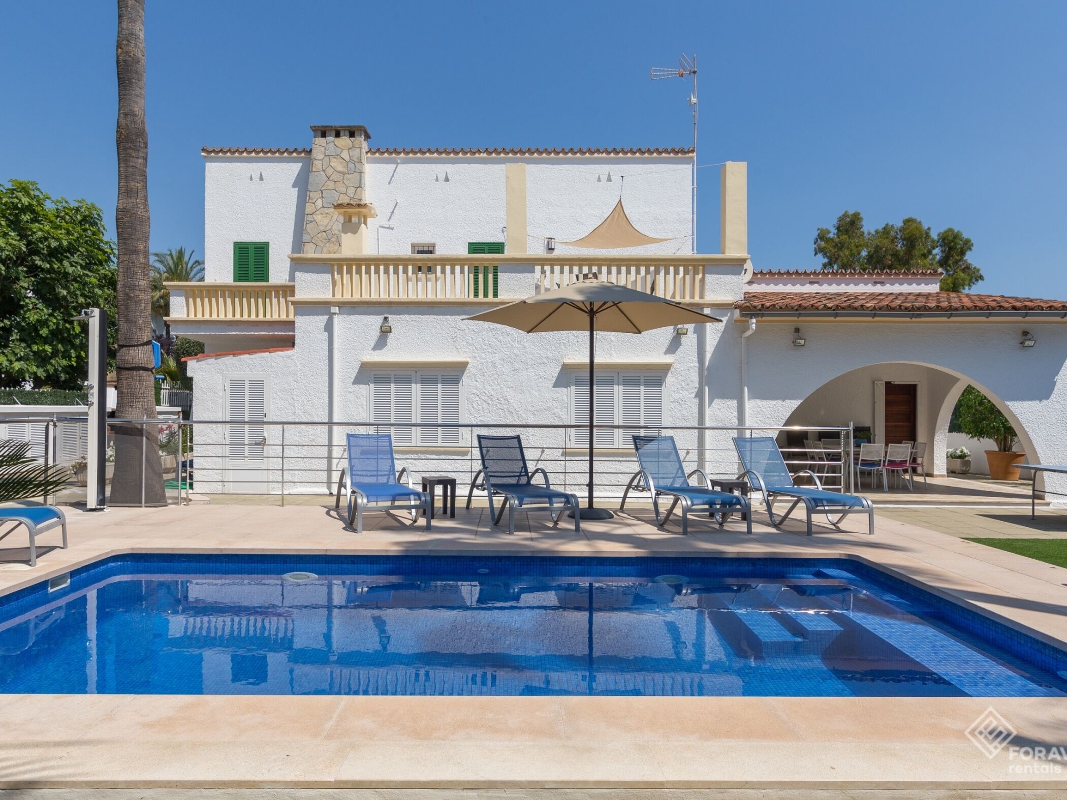 Un área de piscina con sillas de descanso y una mesa, un edificio blanco con techo de tejas y una palmera.