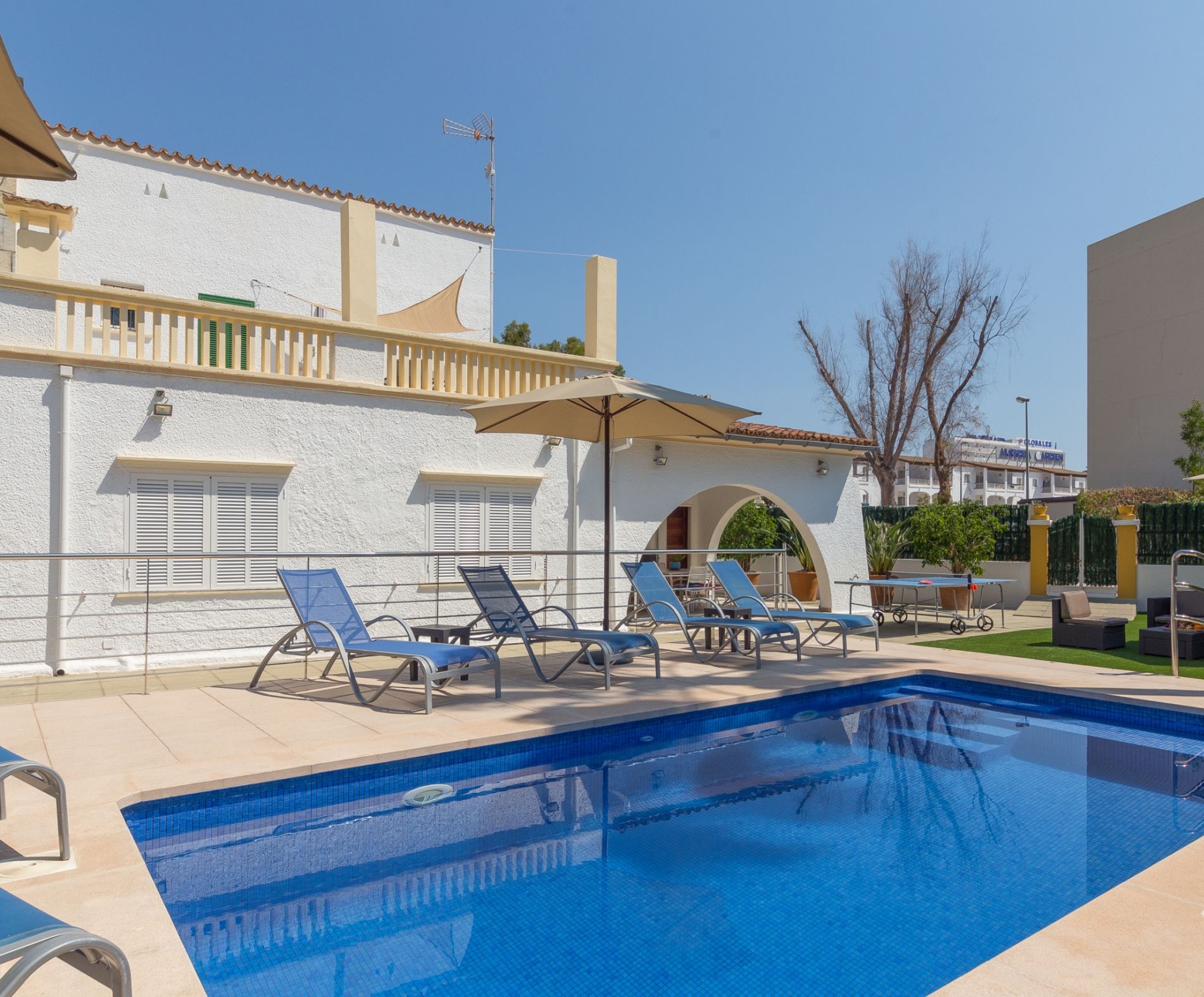 A swimming pool with lounge chairs and umbrellas, surrounded by buildings with balconies and a clear sky.