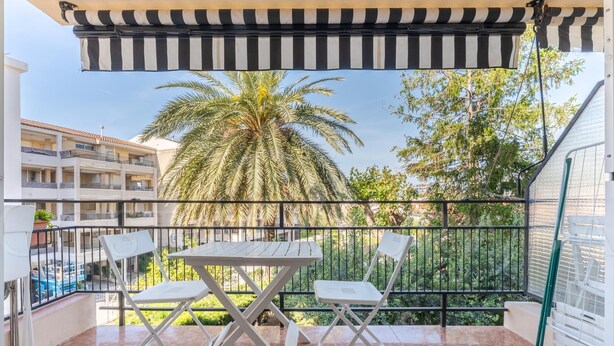 A balcony with a striped awning, two white chairs, a small table, and a view of palm trees and buildings. - 131-POMPEYO II. Apartment located in Cambrils only 300m from the beach (Cambrils)