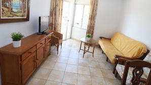 Un salon avec un canapé, un téléviseur posé sur un meuble en bois, une petite table avec une plante et un tableau accroché au mur.