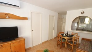 A compact living space with a TV on a wooden cabinet, a dining table with chairs, a small potted plant, and a kitchenette area.