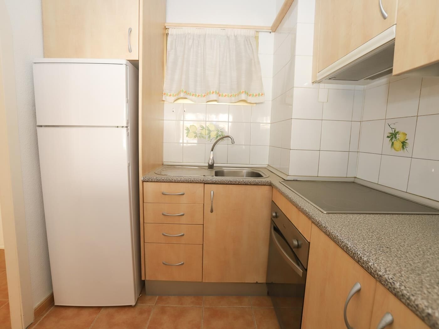 Cocina compacta con refrigerador blanco, armarios de madera, un lavamanos y una ventana con cortina blanca.