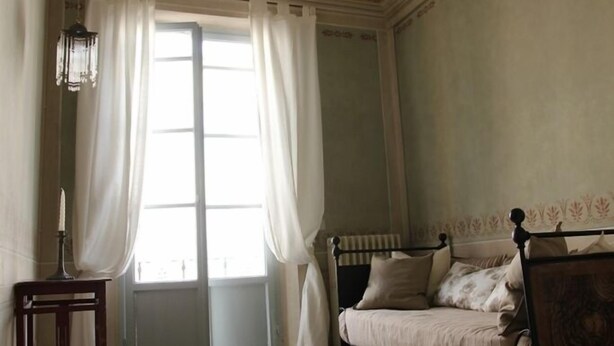Una habitació amb un terra de pissos, un llit amb capçalera de fusta, una taula petita i una finestra amb cortines.