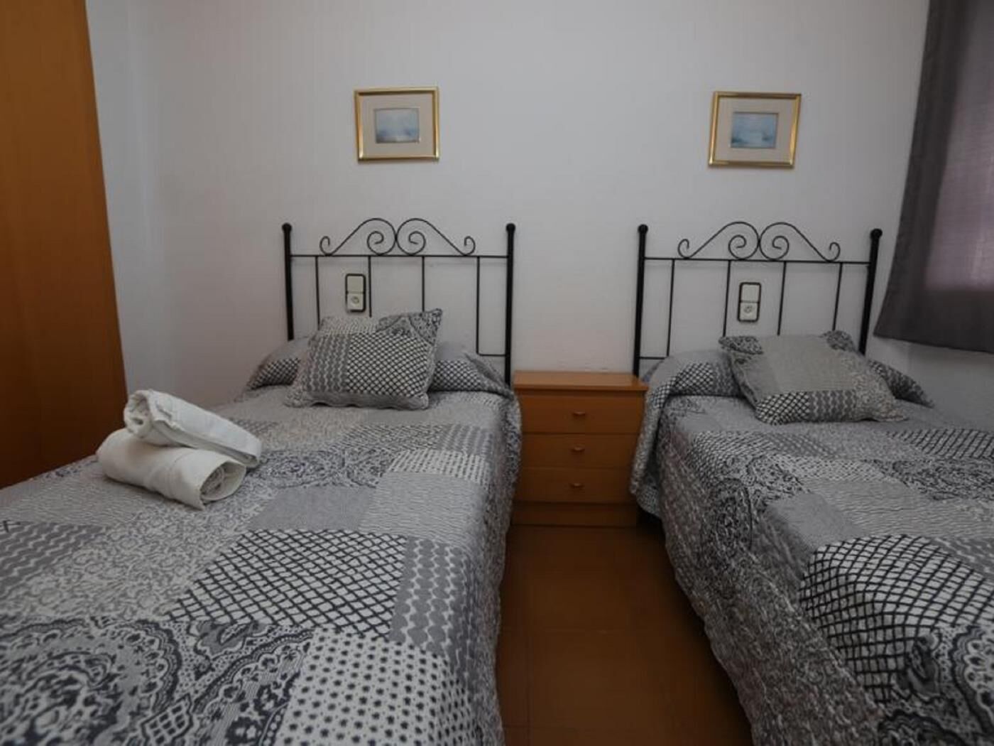 Une chambre avec deux lits, chacun agrémenté de draps et d’oreillers à motifs, une table de chevet en bois et des tableaux encadrés accrochés au mur.