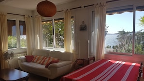 A room with a sofa, a table, a bed with a red bedspread, a fan, and large windows with curtains. - GROUND FLOOR OF VILLA facing the tip of Giens (Carqueiranne)