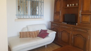 A living room with a white sofa, a wooden cabinet with a television, and a window with blue metal bars. - GROUND FLOOR OF VILLA facing the tip of Giens (Carqueiranne)