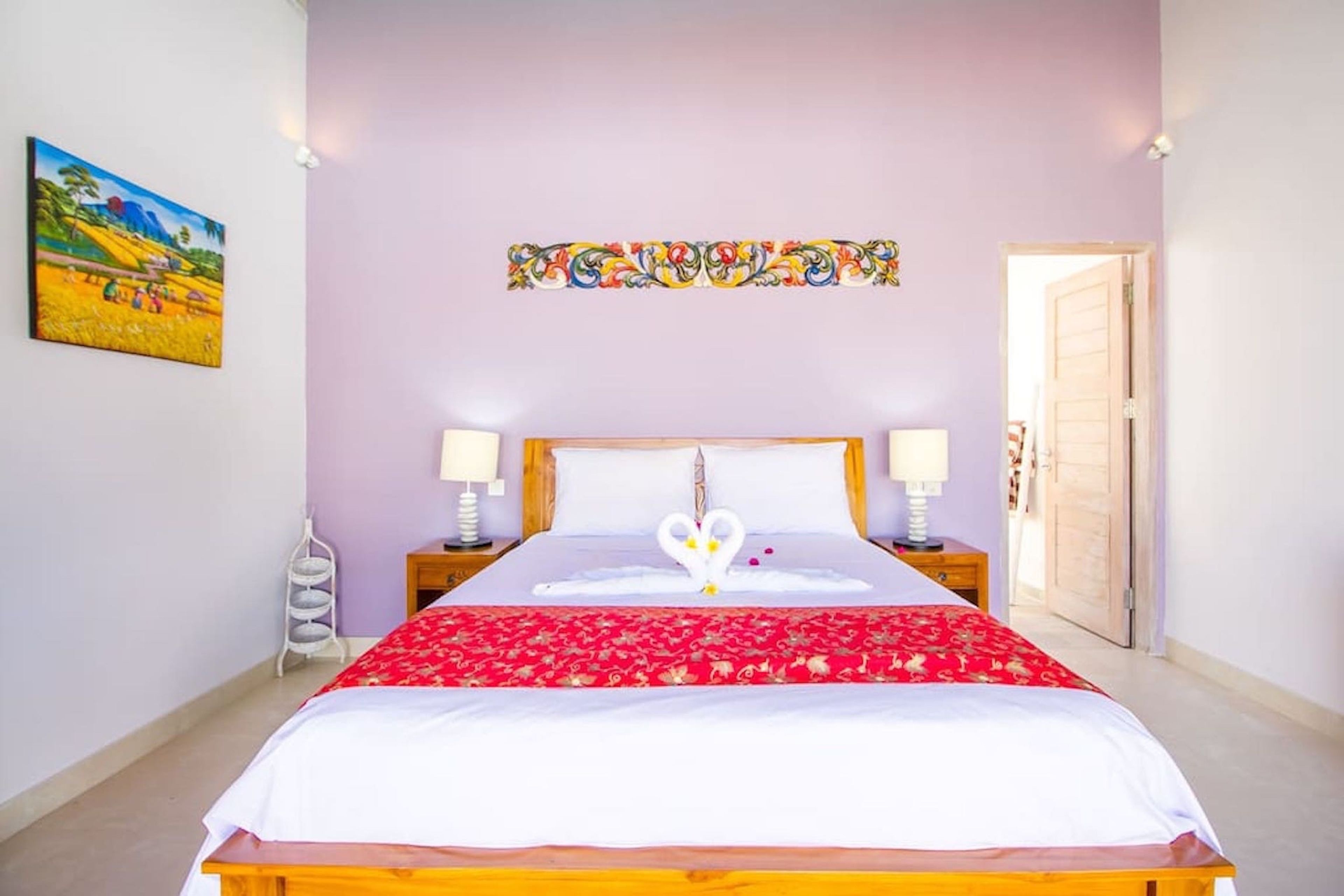 A neatly made bed with a red and white bedspread, two bedside lamps, a decorative wall piece, and a door to the right.