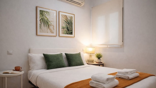 Una cama bien hecha con almohadas blancas y verdes, un edredón marrón y una pila de toallas blancas. Hay una mesita de noche pequeña con una lámpara, una planta en maceta y una taza de café. Encima de la cama, dos láminas enmarcadas con motivos botánicos.