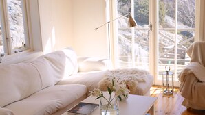 Ein helles Zimmer mit einem weißen Sofa, einem Couchtisch mit Blumenvase und Blick auf eine gebirgige Landschaft durch große Fenster.