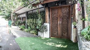 Une entrée d’immeuble avec une porte en bois, de l’herbe artificielle verte et diverses plantes.