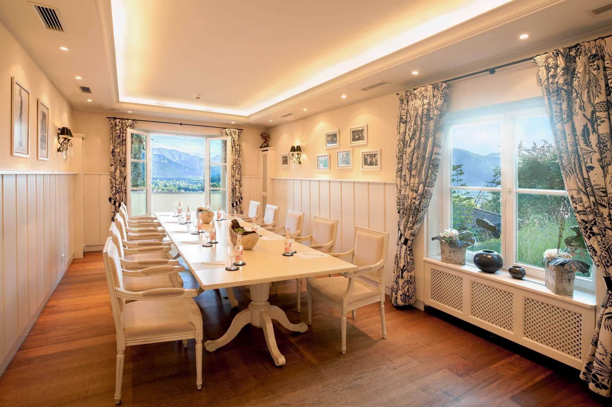 Une salle à manger avec une longue table, des chaises blanches et une vue sur les montagnes, visible  à travers de grandes fenêtres.