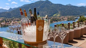 A glass table with a wine cooler containing several bottles and glasses, set against a scenic mountain backdrop. - Suite - Hotel Leeberghof (Tegernsee)