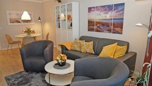Een moderne woonkamer met een grijze bank, twee fauteuils, een salontafel met een bloevas, een boekenkast en een schilderij van een zonsondergang boven de oceaan.