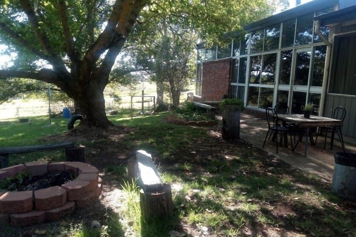 A patio area with a brick fire pit, a wooden table with chairs, and a large tree providing shade.