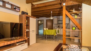 Un vaste espace de vie doté d’un plafond en bois, d’un grand téléviseur à écran plat, d’une table à manger avec des chaises, et d’une cuisine située au fond de la pièce.