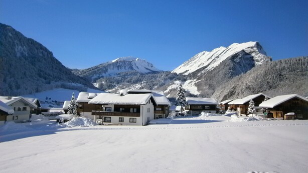 A snow-covered mountain village with chalet-style buildings, a clear blue sky, and a prominent mountain peak in the background.