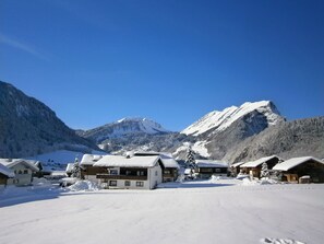 Un village de montagne enneigé, avec des bâtiments de style chalet, un ciel bleu dégagé et un sommet montagneux imposant en arrière-plan.