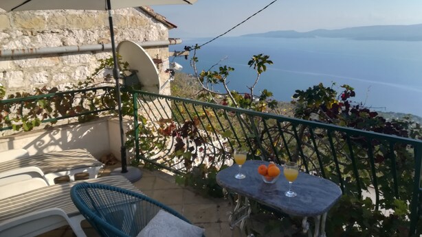 A balcony with a table, chairs, and an umbrella overlooking a scenic view. - Charming stone house with panoramic sea and mountain view (Lokva Rogoznica)