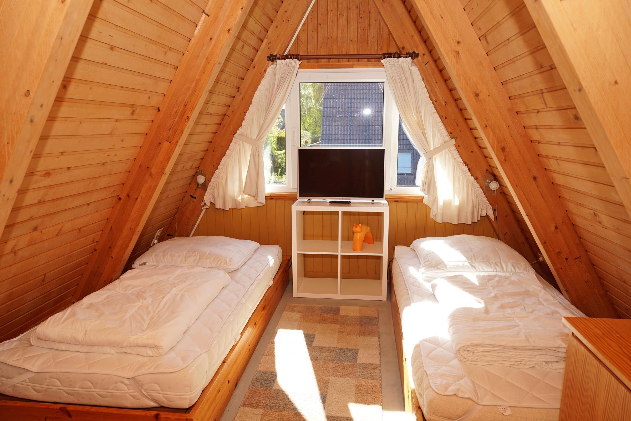 A small, cozy attic bedroom with two beds, a TV, and a window with curtains.