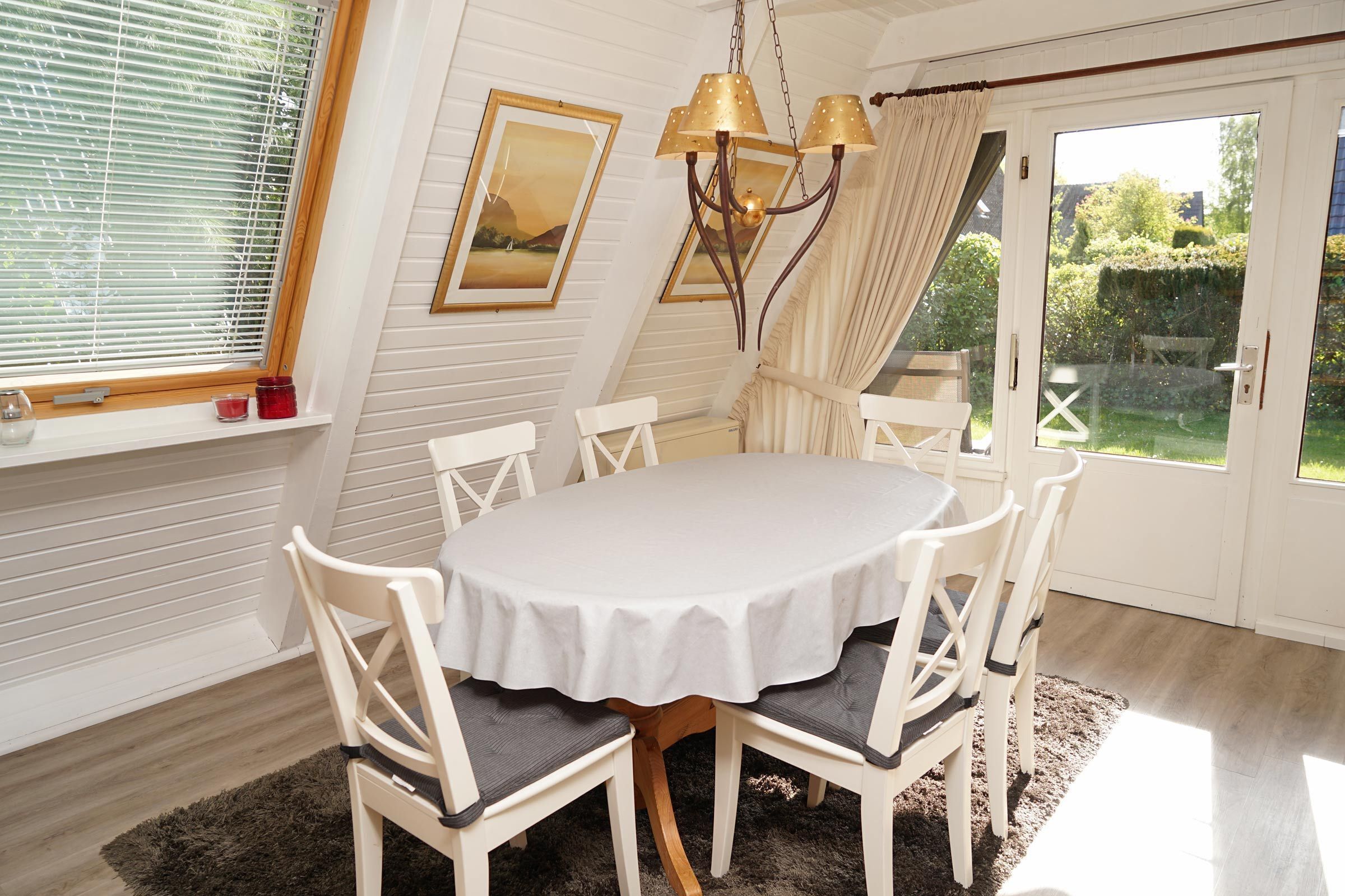 A dining area with a round table, white chairs, a chandelier, and a view of the garden through a glass door.
