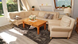Ein gemütliches Wohnzimmer mit einem beigefarbenen Sofagarnitur, einem Holztisch und einem großen Fenster mit Jalousien.