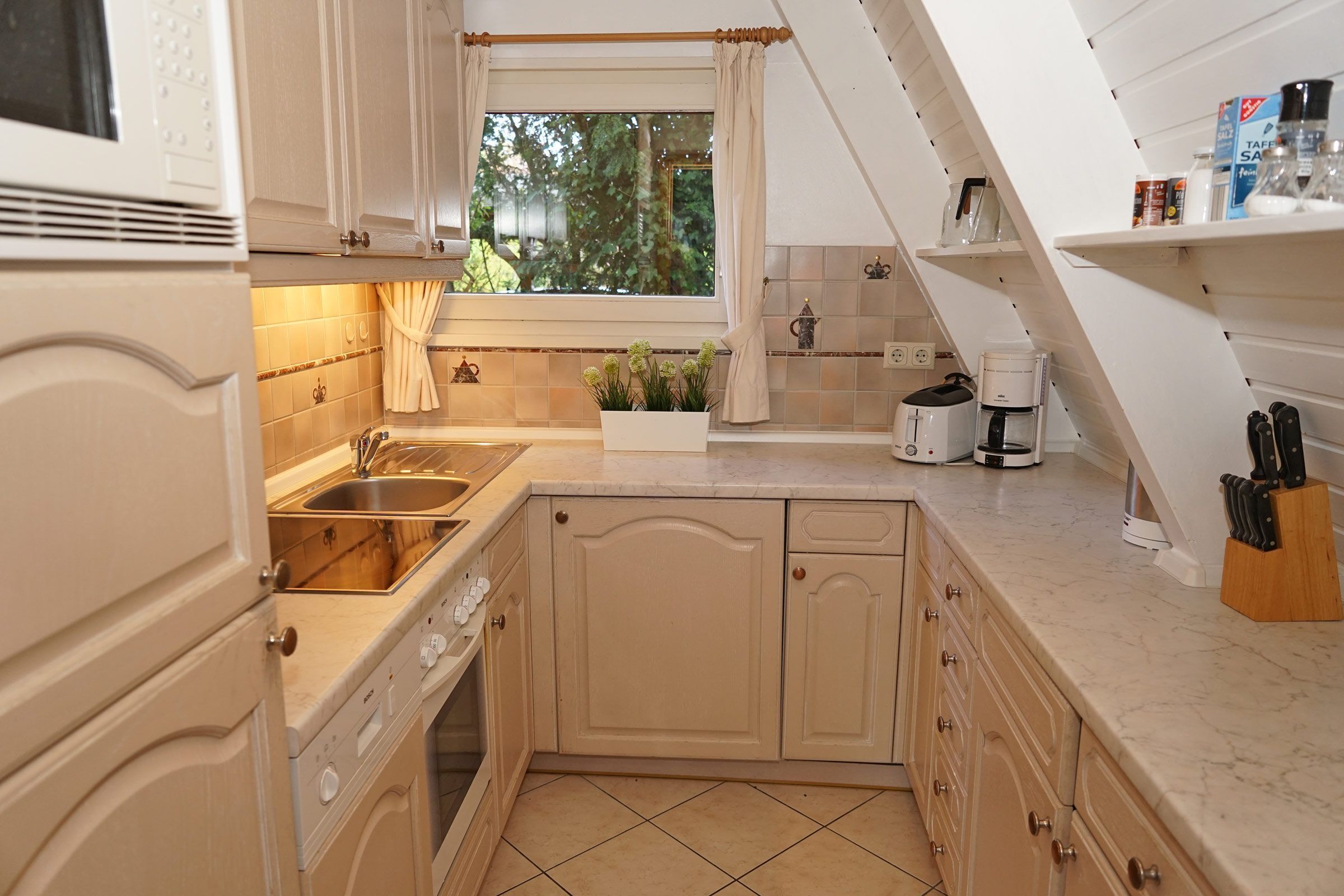 Una cocina con pileta, estufa y cafetera. Hay una ventana con cortinas y una planta sobre la encimera.