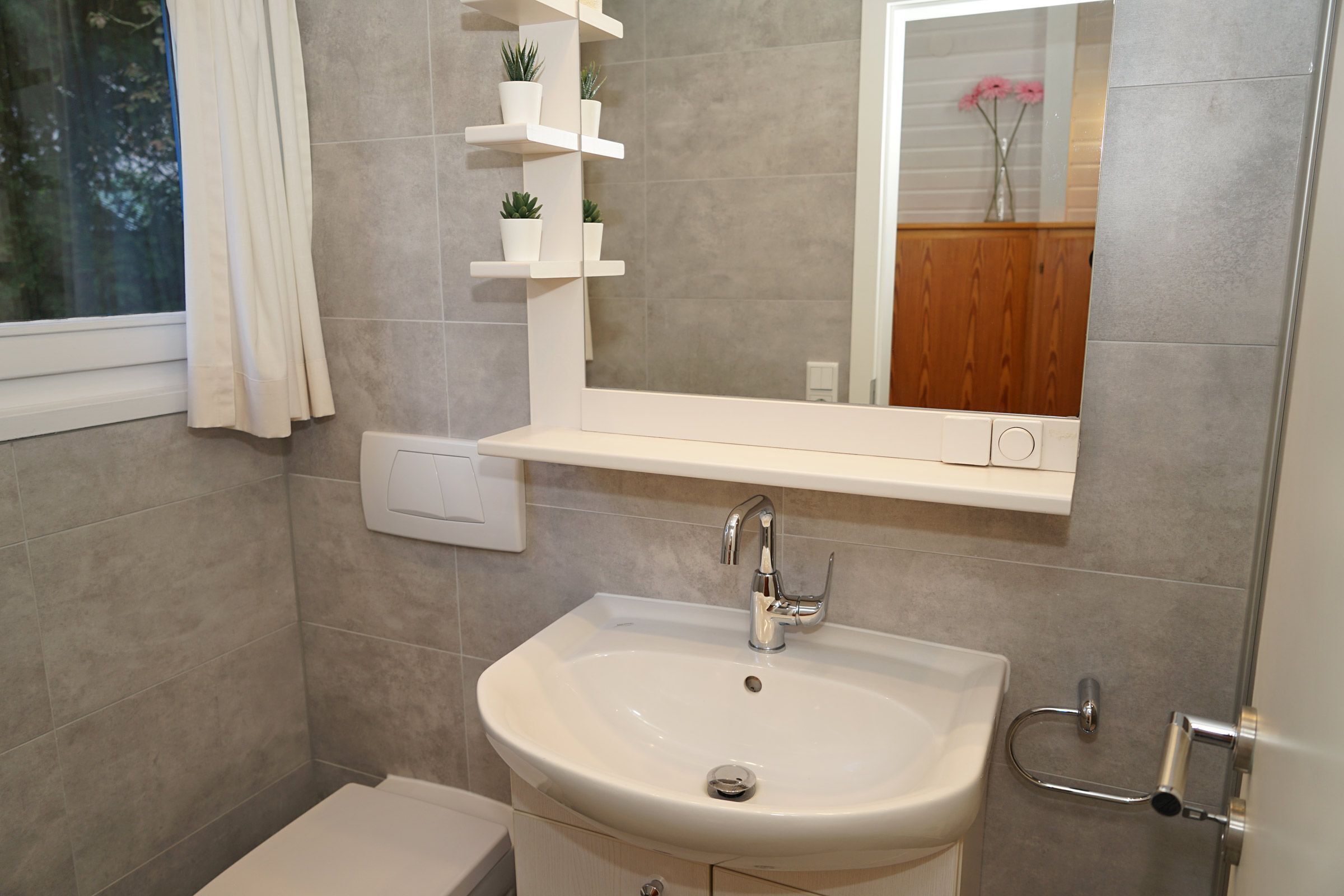 Casa de banho com lavatório branco, espelho e prateleira com plantas em vasos.