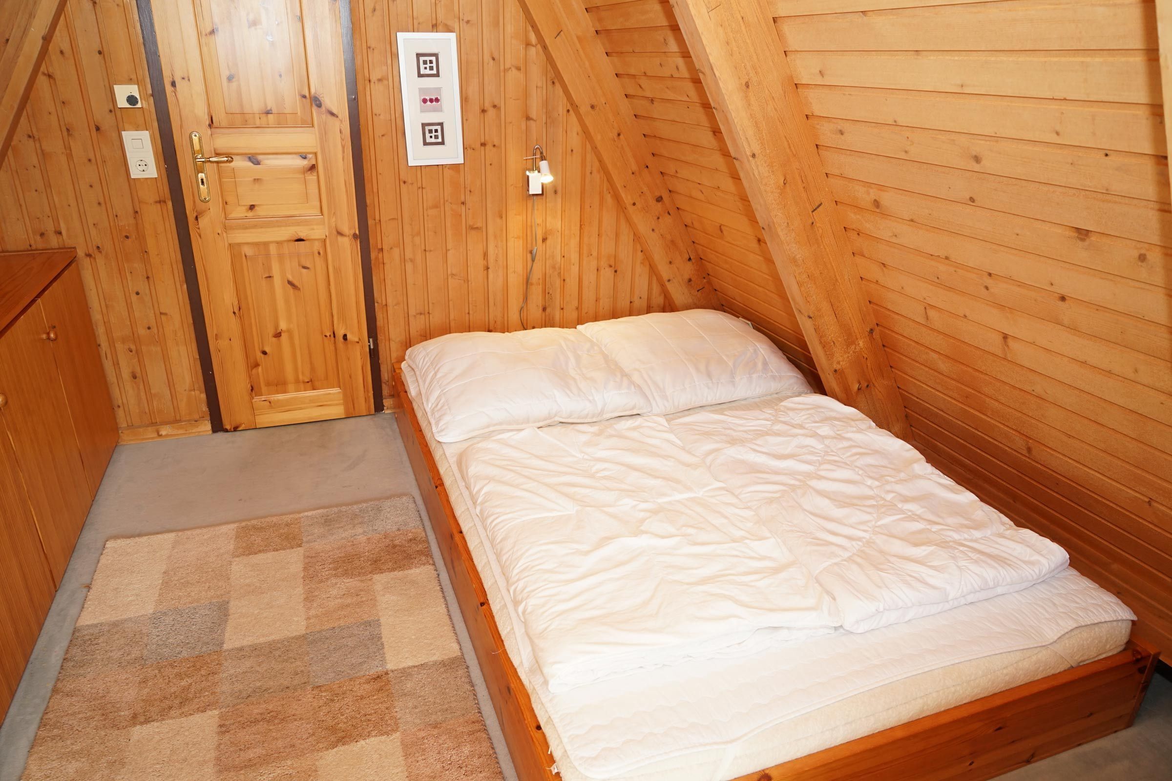 Una habitación con paneles de madera, una cama individual, una puerta y un pequeño aplique en la pared.